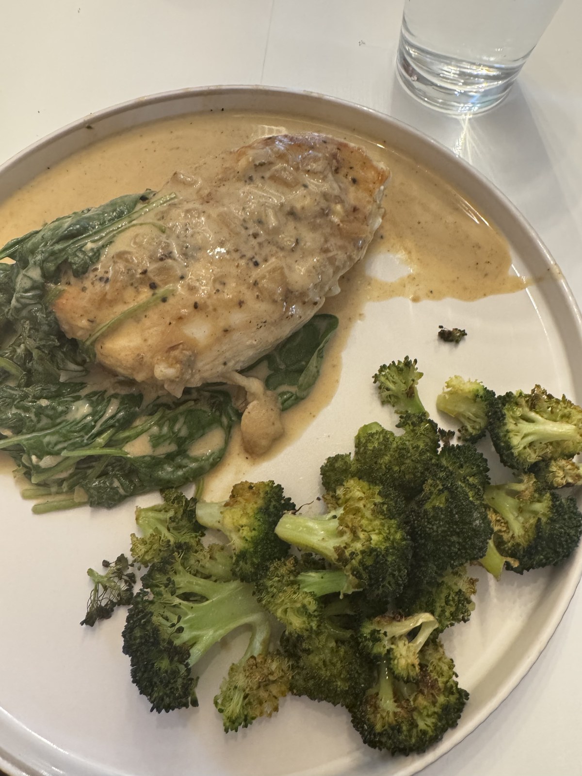 Chicken breast under a Dijon cream sauce on a bed of creamed spinach, with roasted broccoli alongside on a white plate