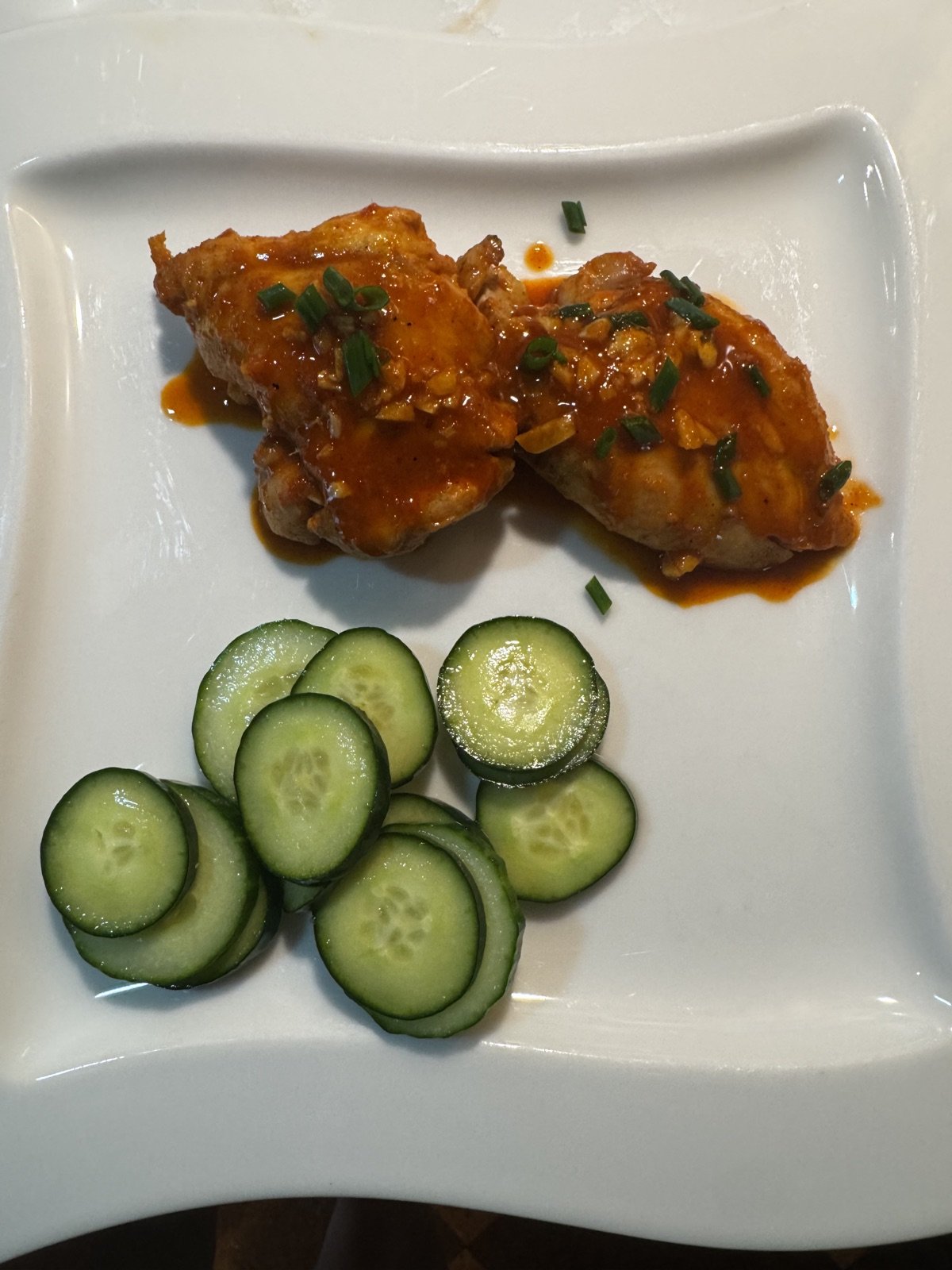 Two gochujang-glazed chicken thighs topped with sliced scallions on a white plate, with rounds of cucumber alongside