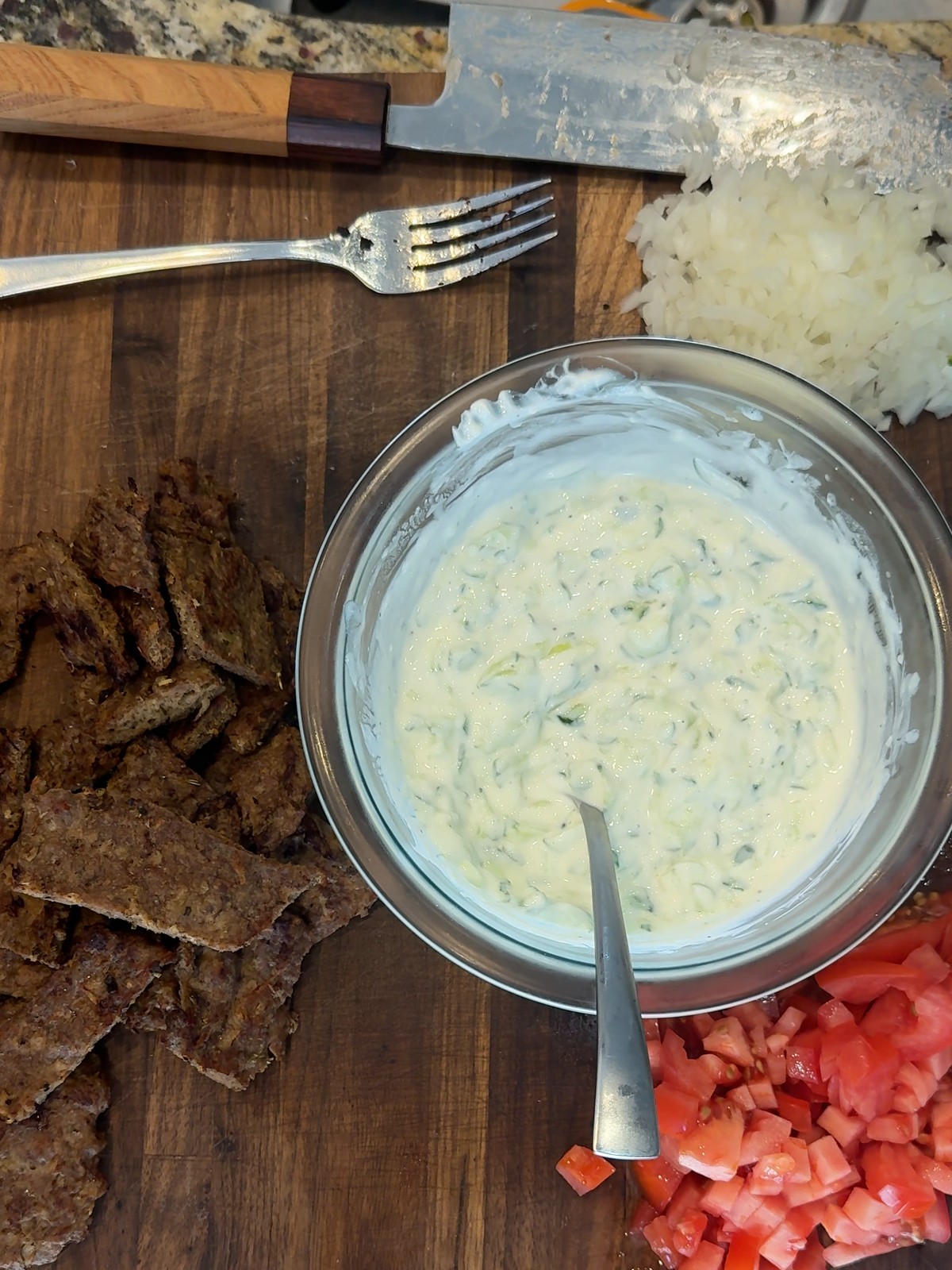 Crisped lamb strips, a bowl of cucumber tzatziki, diced tomato, and finely chopped onion laid out on a wood board