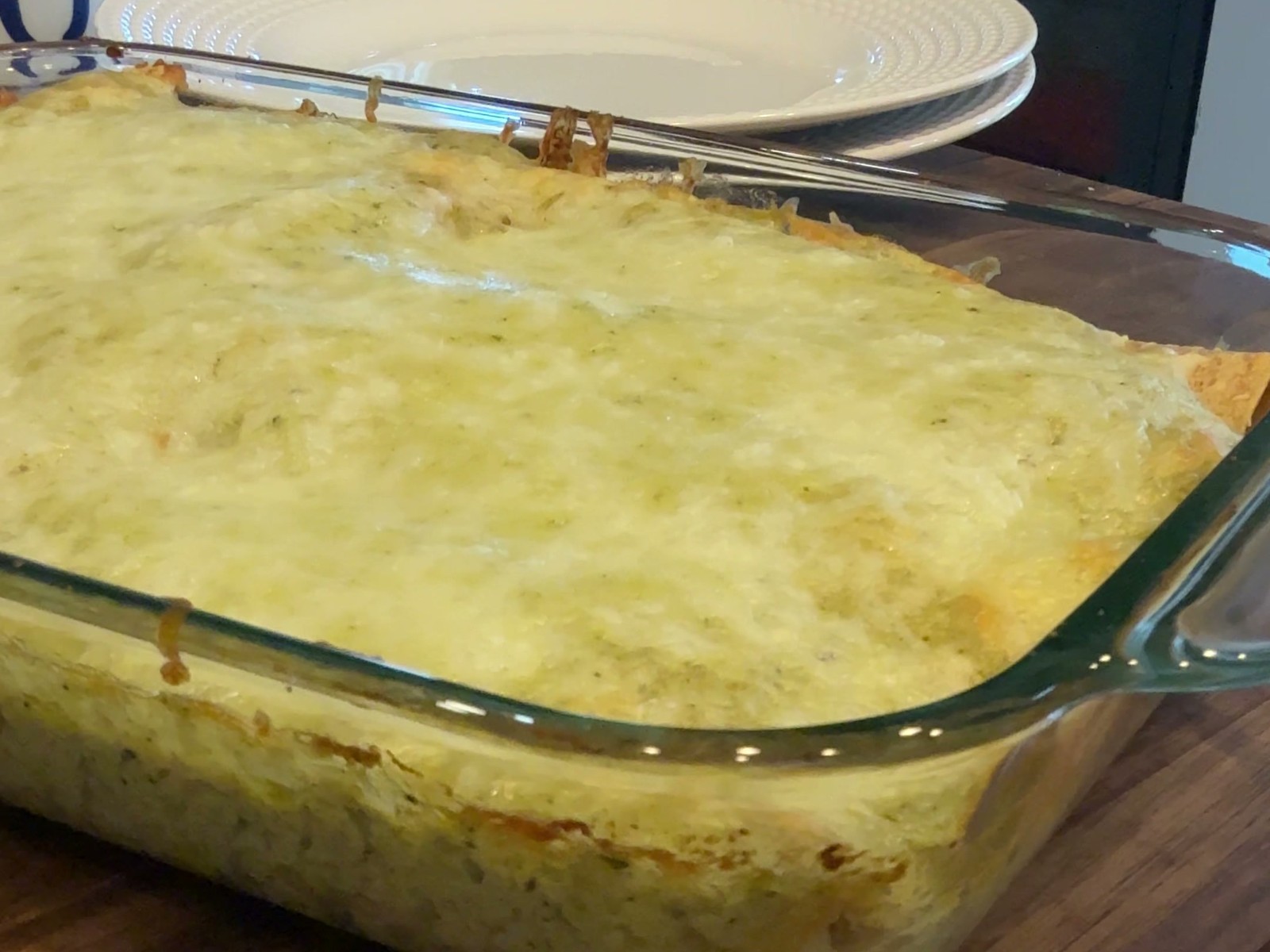 A glass baking dish of chicken enchiladas baked under pale green poblano cream and melted queso fresco, browned at the edges, resting on a walnut board