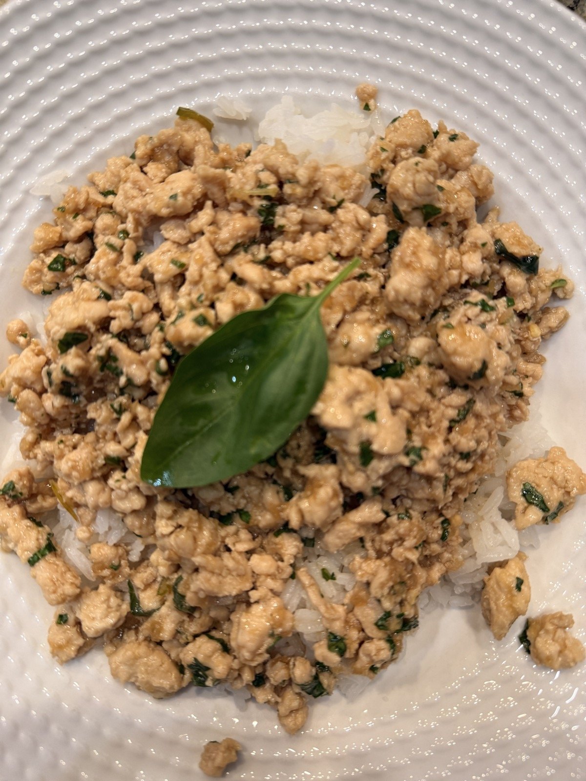 Ground chicken cooked with chopped basil in a glossy sauce, spooned over white rice on a white plate with a whole basil leaf on top